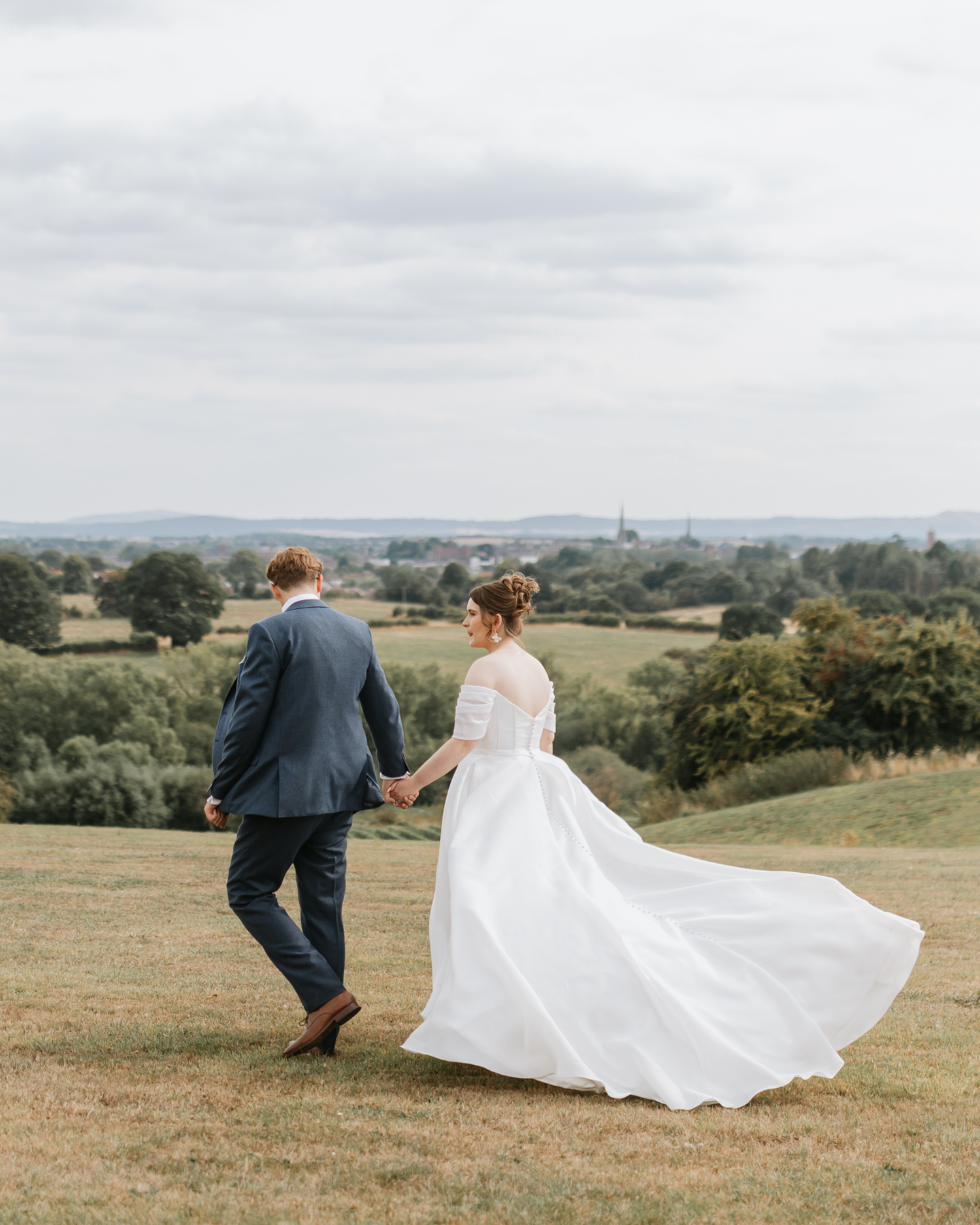 A couple in wedding attire walk hand in hand across a scenic hilltop meadow with rolling countryside views in the background.