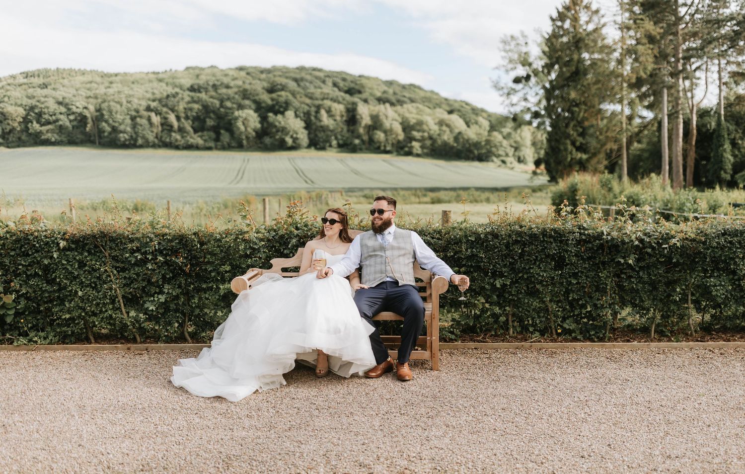 A couple in wedding attire lying on a gravel path with green trees and countryside in the background.