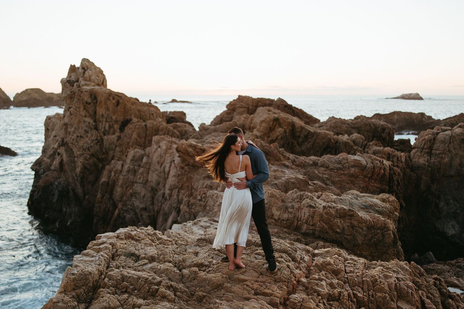 A romantic couple stands on rocky coastal cliffs at sunset overlooking the ocean.