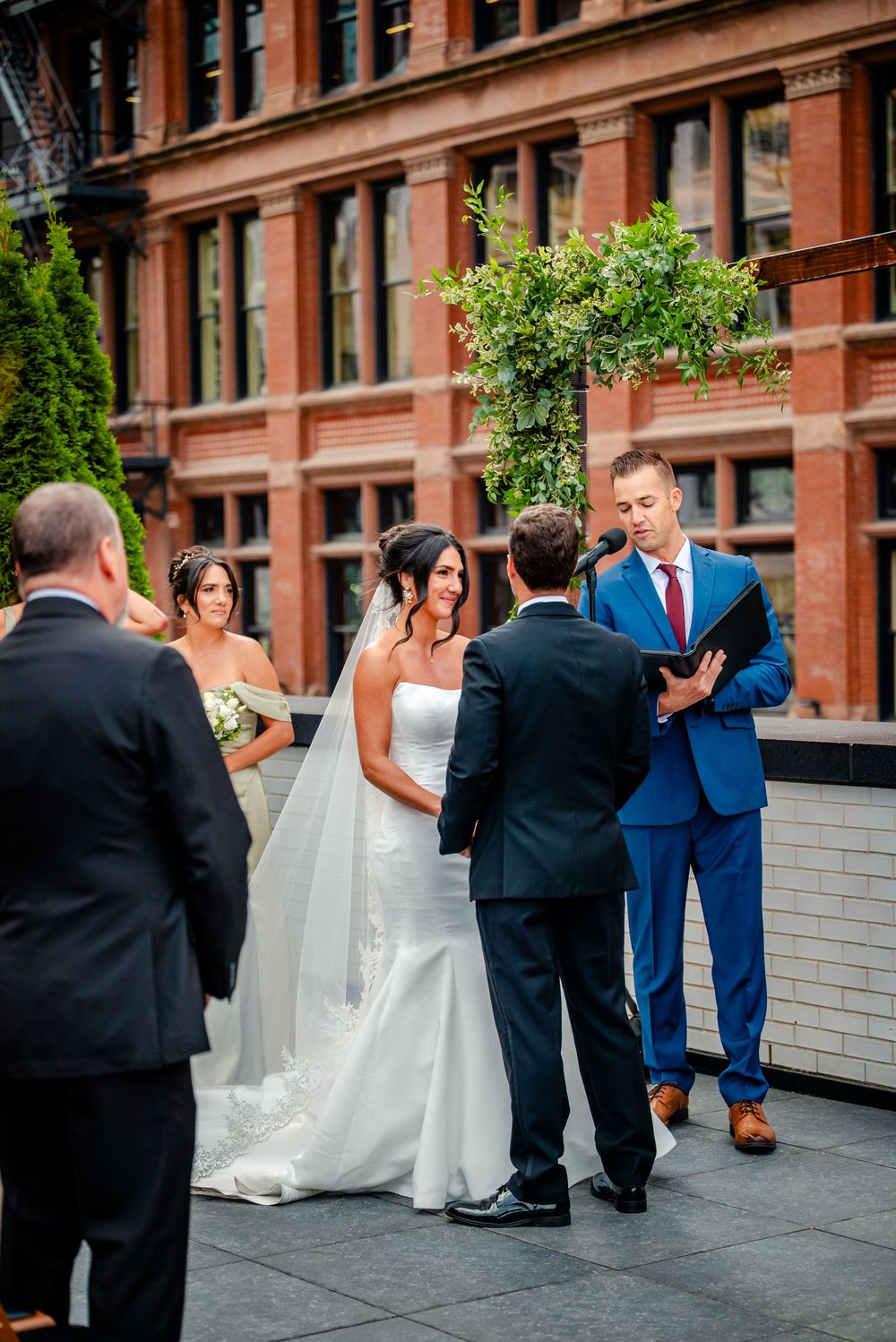 An outdoor wedding ceremony takes place in front of a historic brick building with guests gathered on a rooftop terrace.