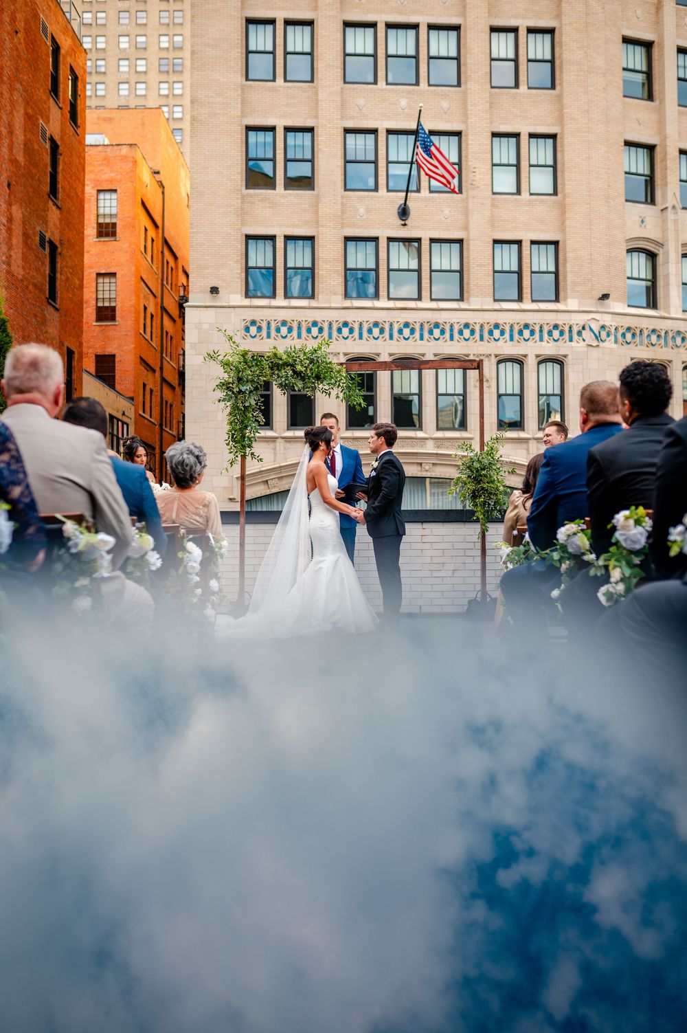 Outdoor rooftop wedding ceremony in urban setting with American flag and historic architecture in background.