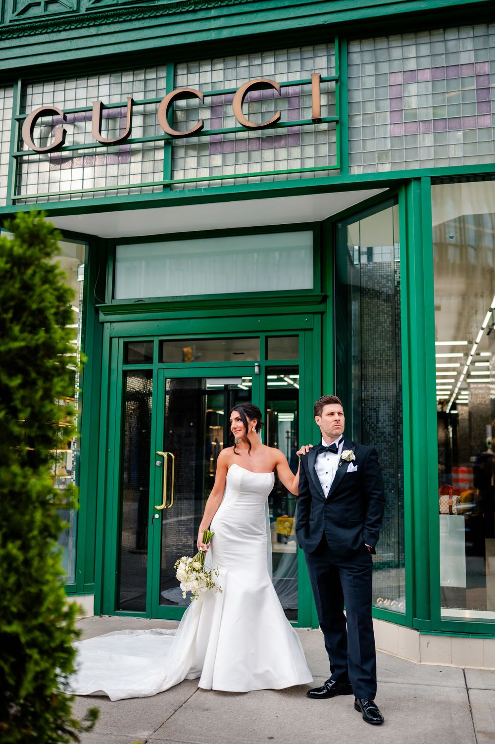 A wedding couple poses in front of a green-trimmed storefront with tiled walls and glass windows.