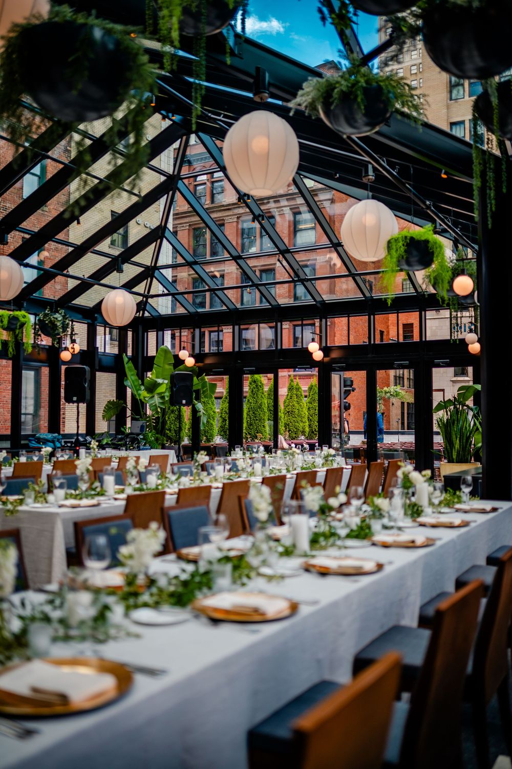 Elegant wedding reception table setting under a glass ceiling conservatory with hanging paper lanterns and greenery.