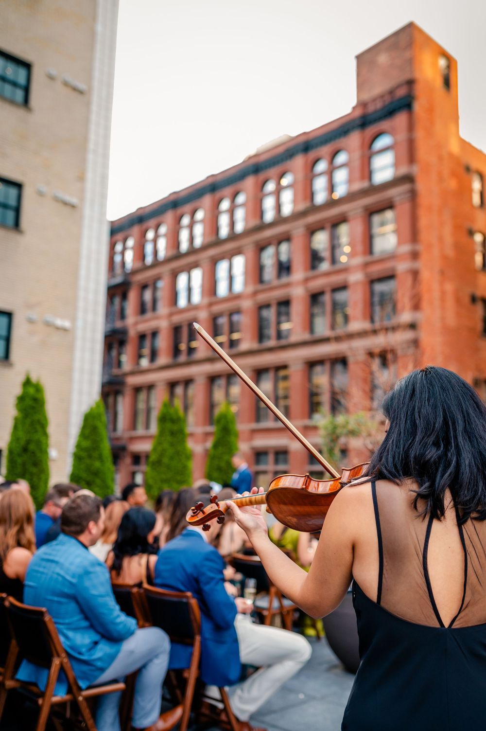 Violinist in black dress performs at outdoor rooftop event with red brick building backdrop and seated audience.