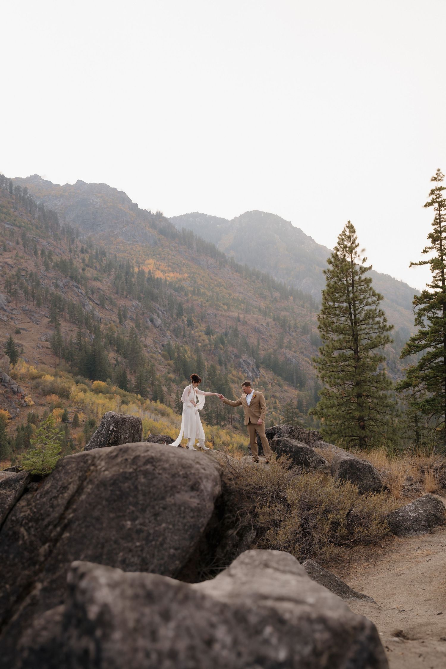 A couple stands together on a rugged mountain cliff with vibrant fall colors and desert landscape in the background. Leavenworth, WA