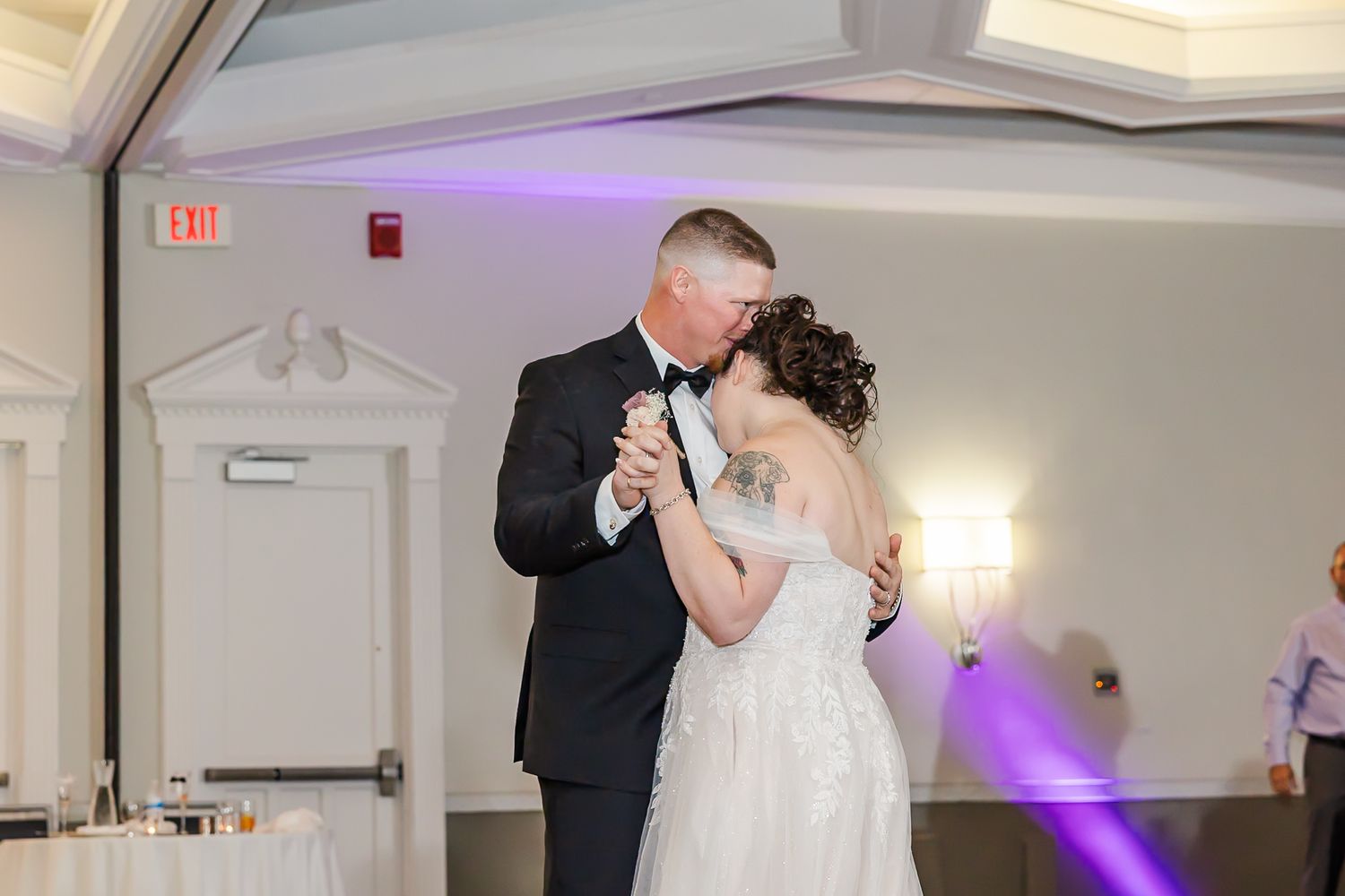 A couple shares a tender moment while dancing together at a wedding reception with purple lighting in the background.