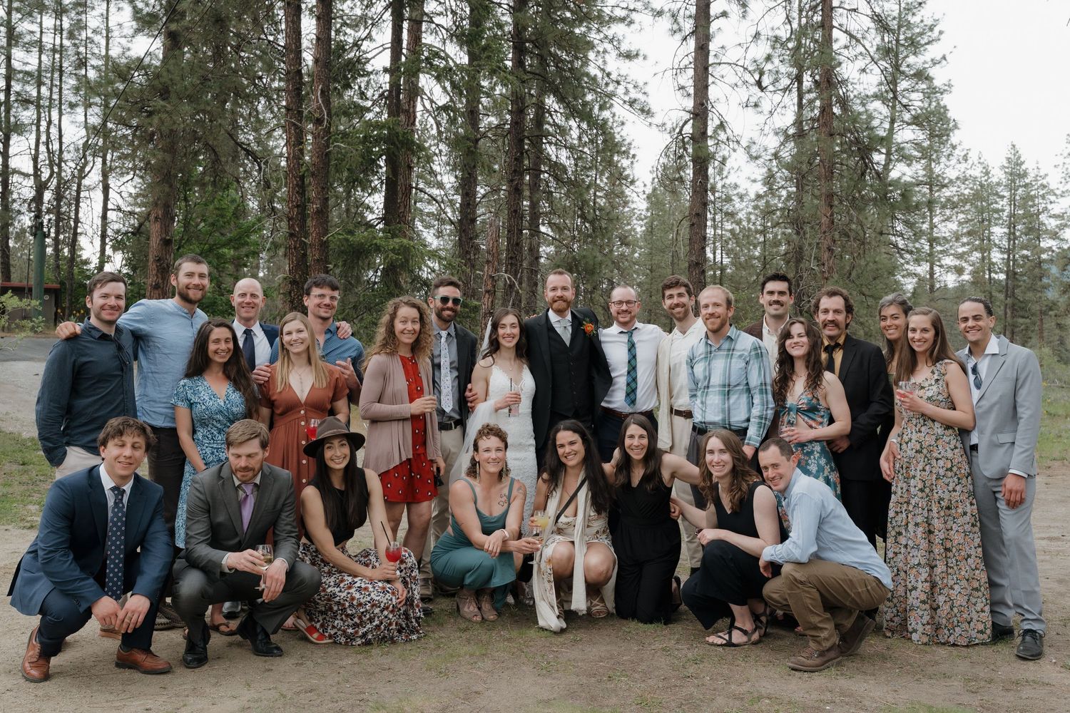 Large group of wedding guests pose together outdoors in a scenic forest setting with tall pine trees.