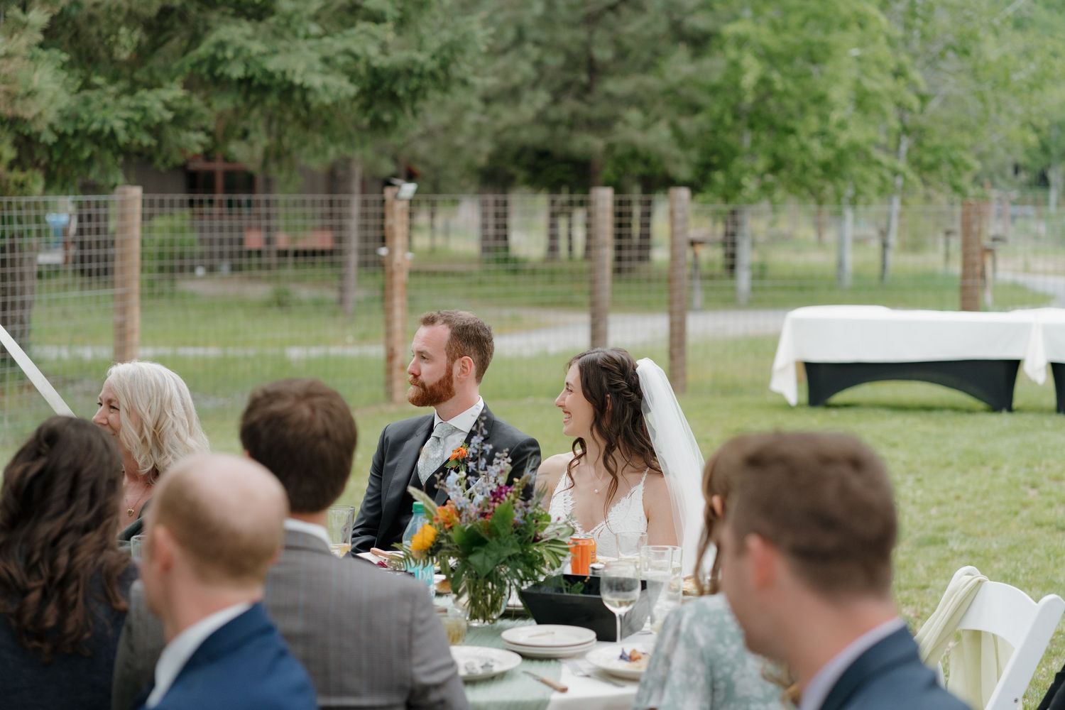 Guests seated at outdoor tables during a casual garden party celebration with white chairs and green foliage.