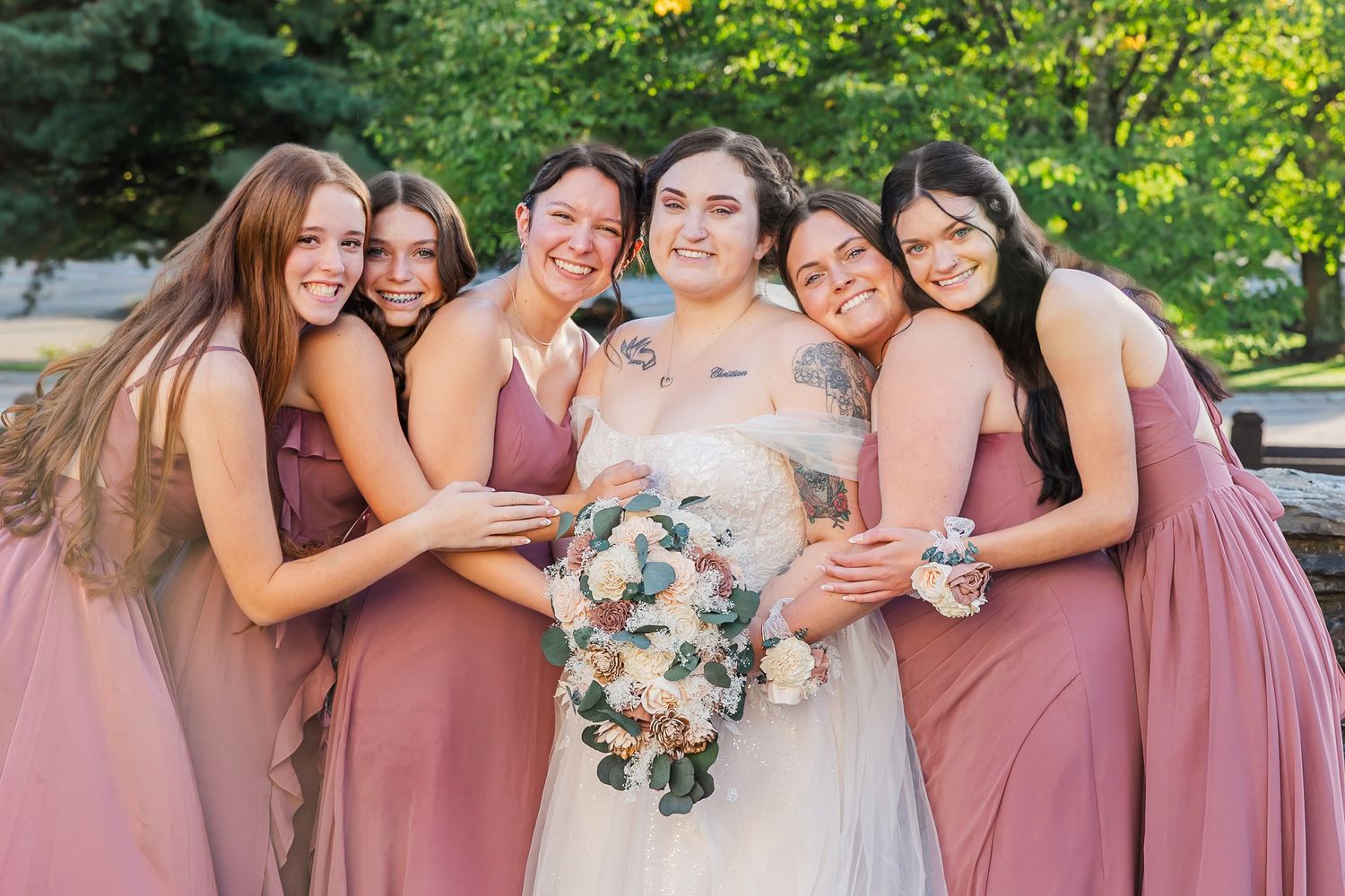 A group in dusty rose bridesmaid dresses and floral bouquets share joyful moments during an outdoor wedding celebration.