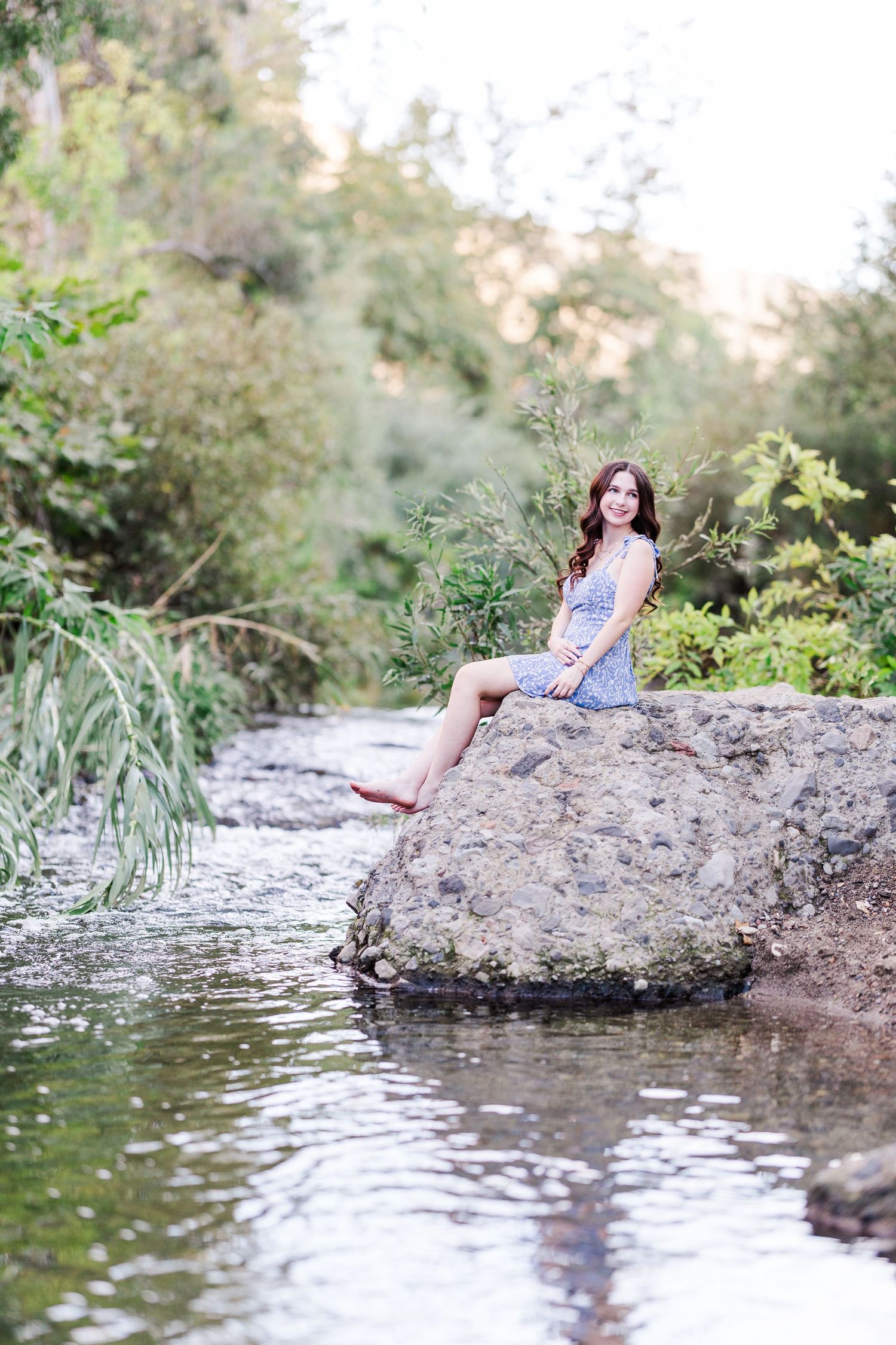 A figure sits peacefully on a large rock formation by a tranquil stream in a natural outdoor setting.