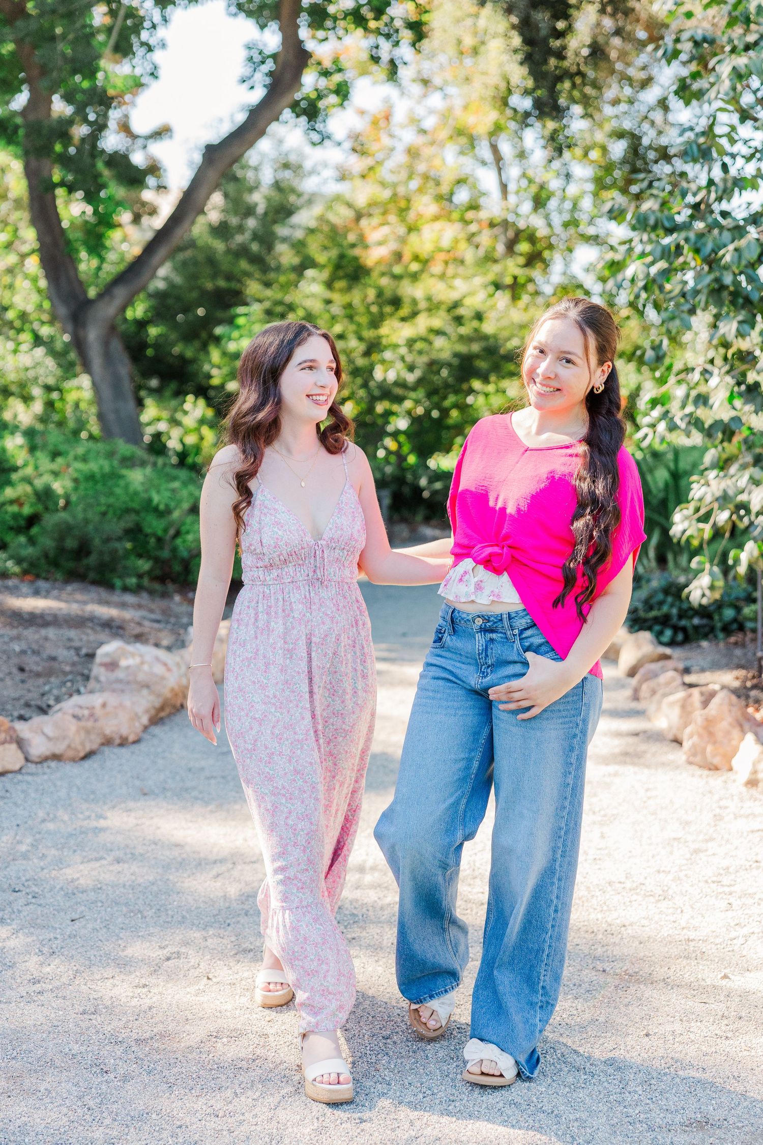 Two friends walking together on a path, one wearing a floral dress and one in jeans and pink sweater on a sunny day.