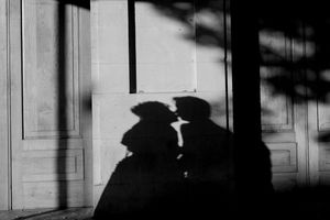 A black and white photograph shows silhouettes of people walking along a sidewalk with dramatic shadows cast on a wall.