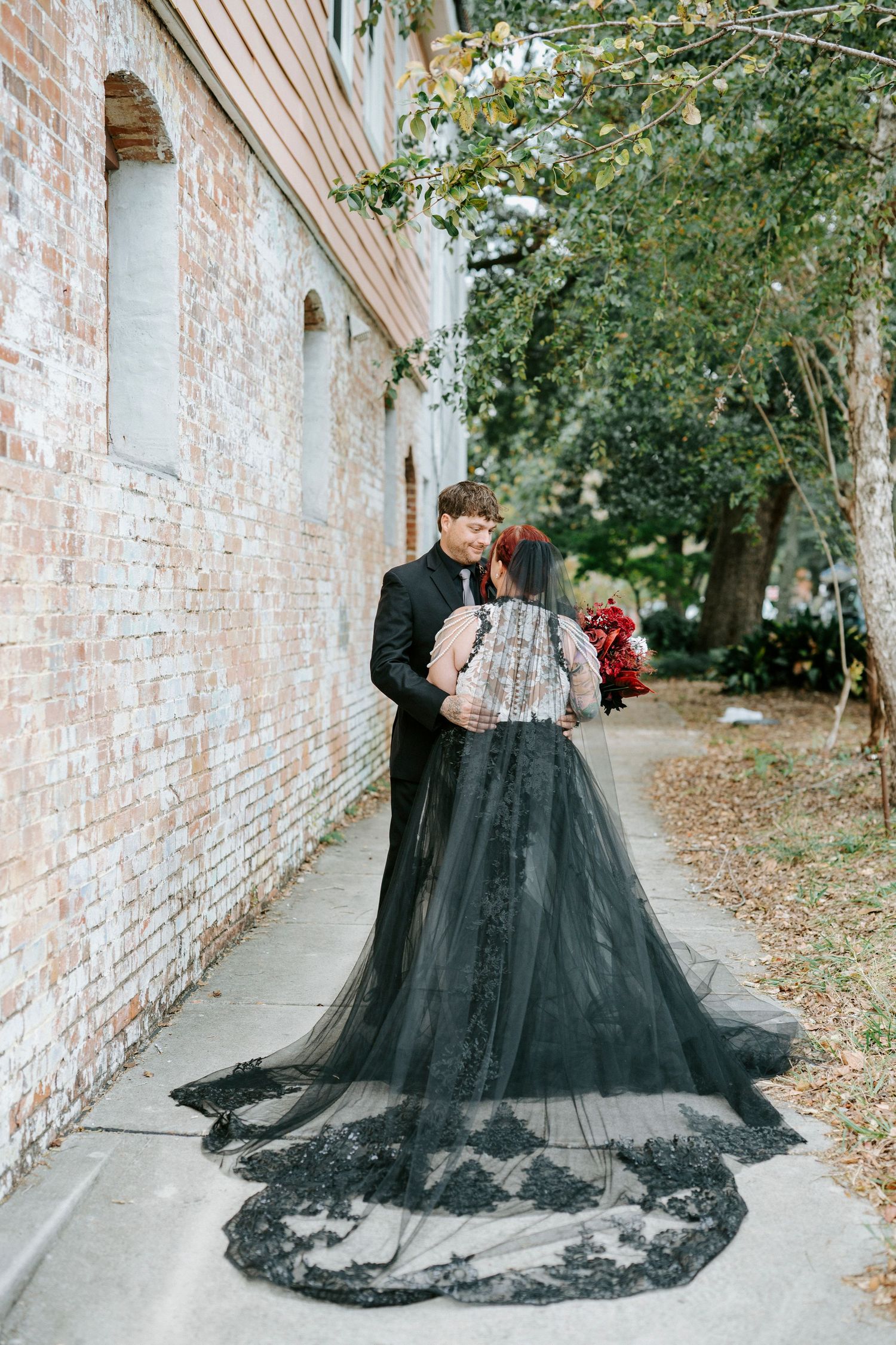 A dramatic black tulle wedding gown with long train photographed against a rustic brick wall outdoors.