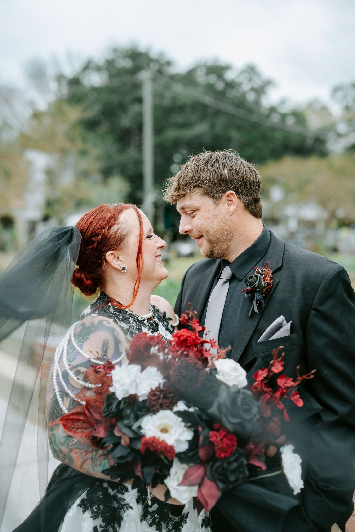 A romantic wedding couple shares an intimate moment with red and white floral bouquet against a cloudy sky.