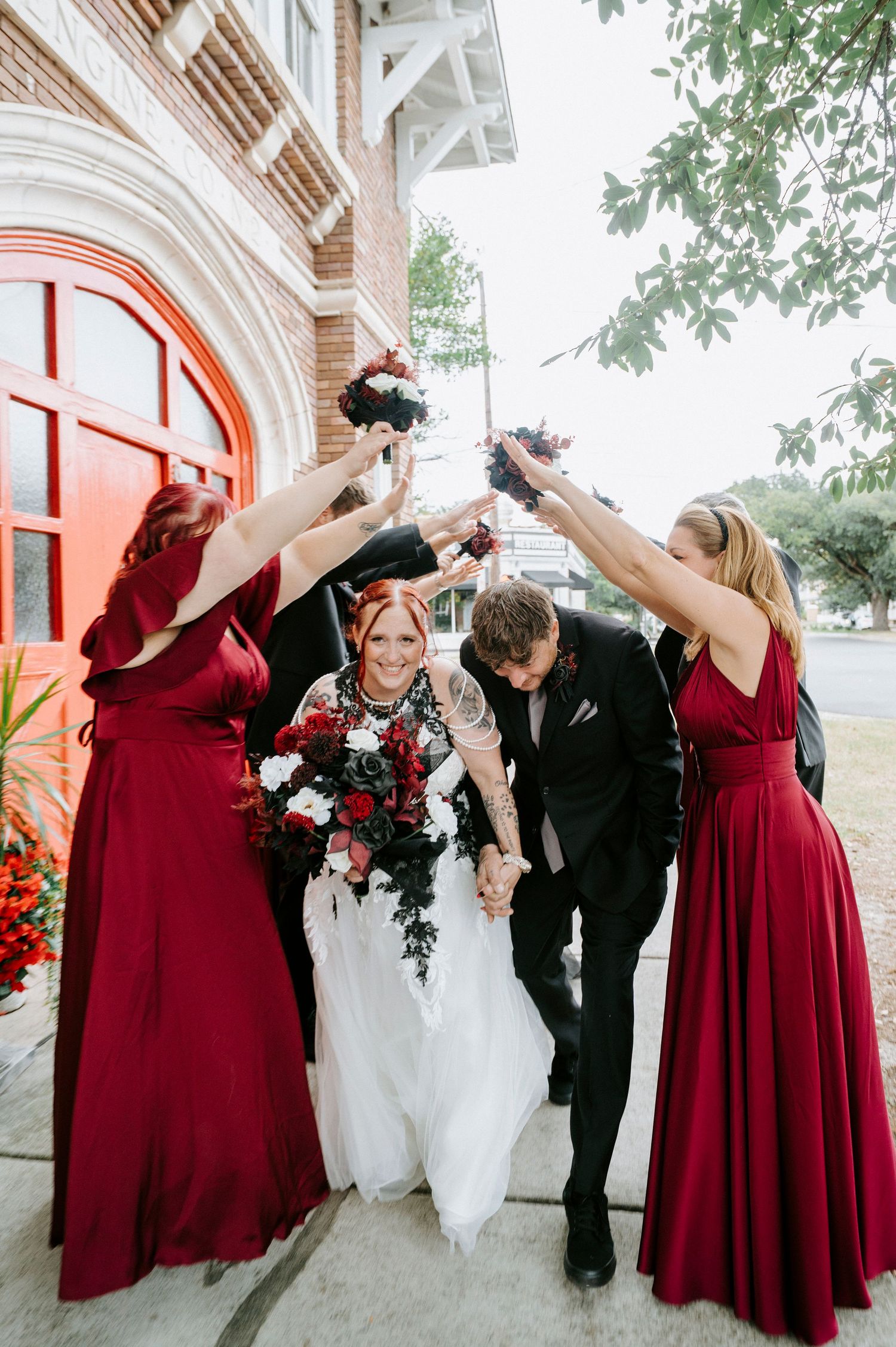 Wedding party in burgundy bridesmaid dresses and black suit celebrate joyfully outside brick building with arched doorway.