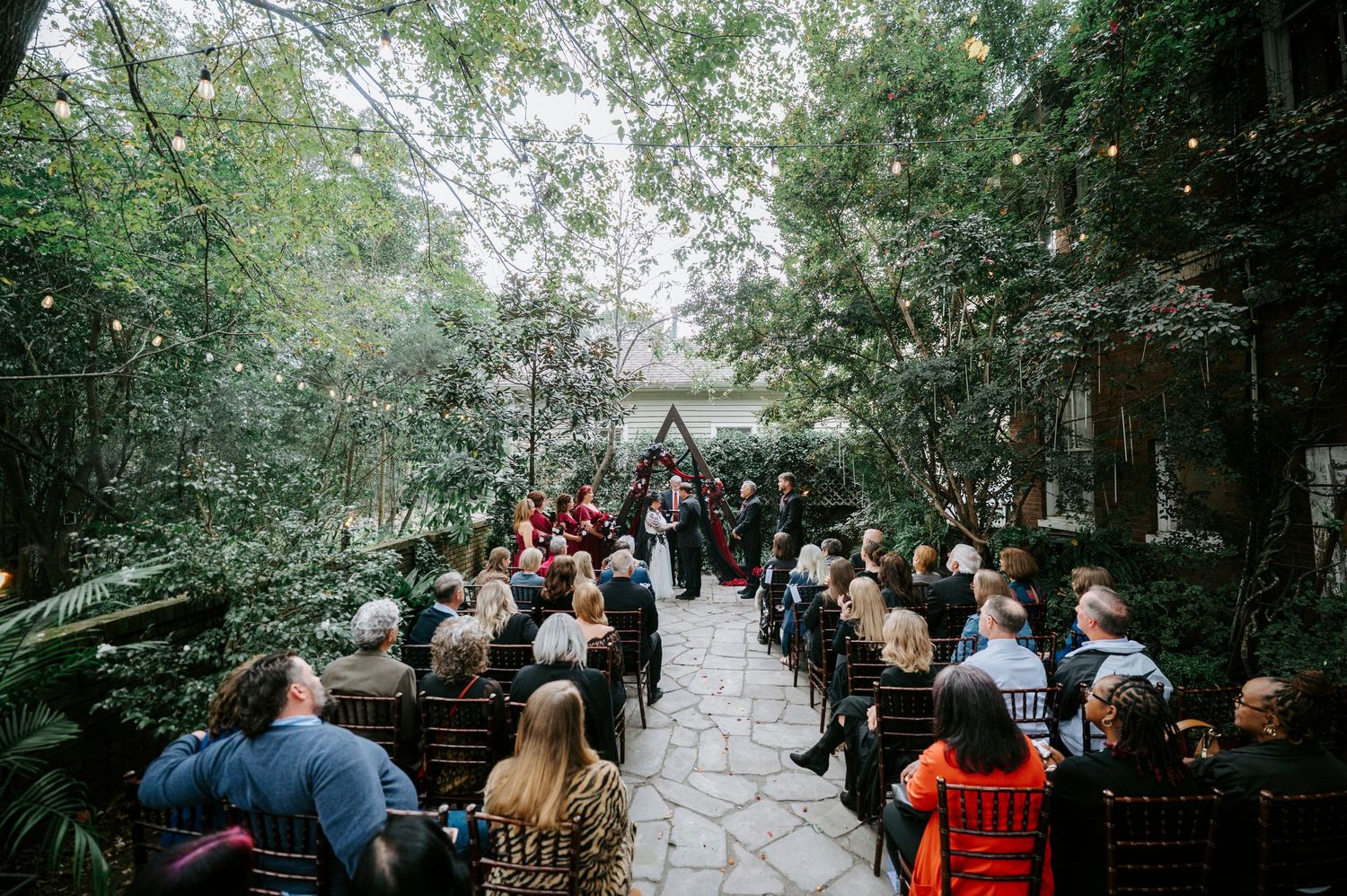 A line of wedding guests walking down a tree-lined stone pathway at an outdoor ceremony venue.