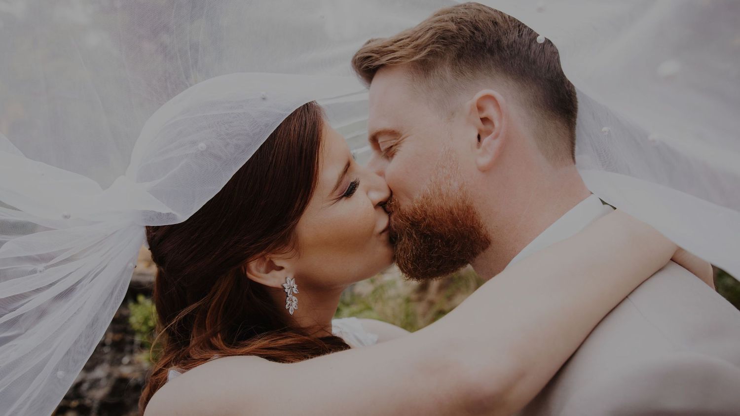Bride and groom sharing romantic kiss under flowing veil in outdoor wedding portrait.