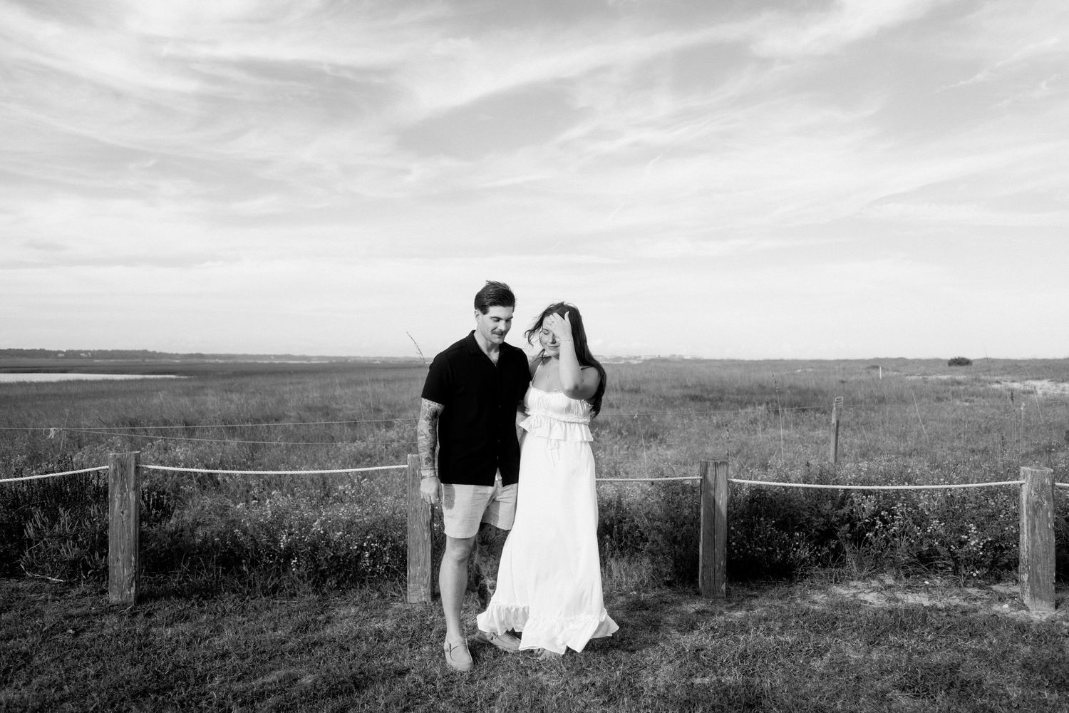 A couple in wedding attire walks through a scenic meadow with mountains in the background during sunset.