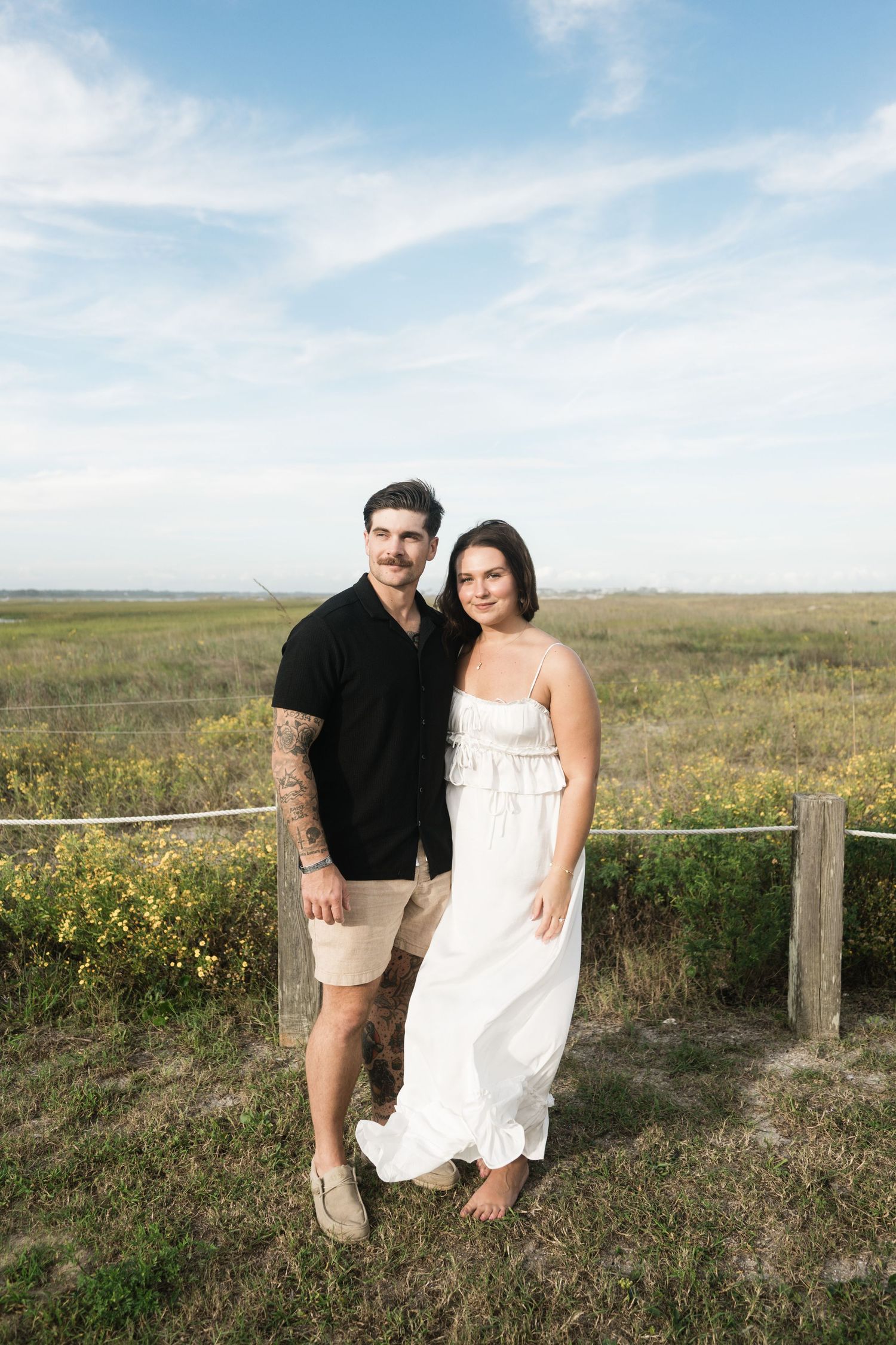 A couple poses together in a scenic prairie field with wildflowers and blue sky on a sunny day.