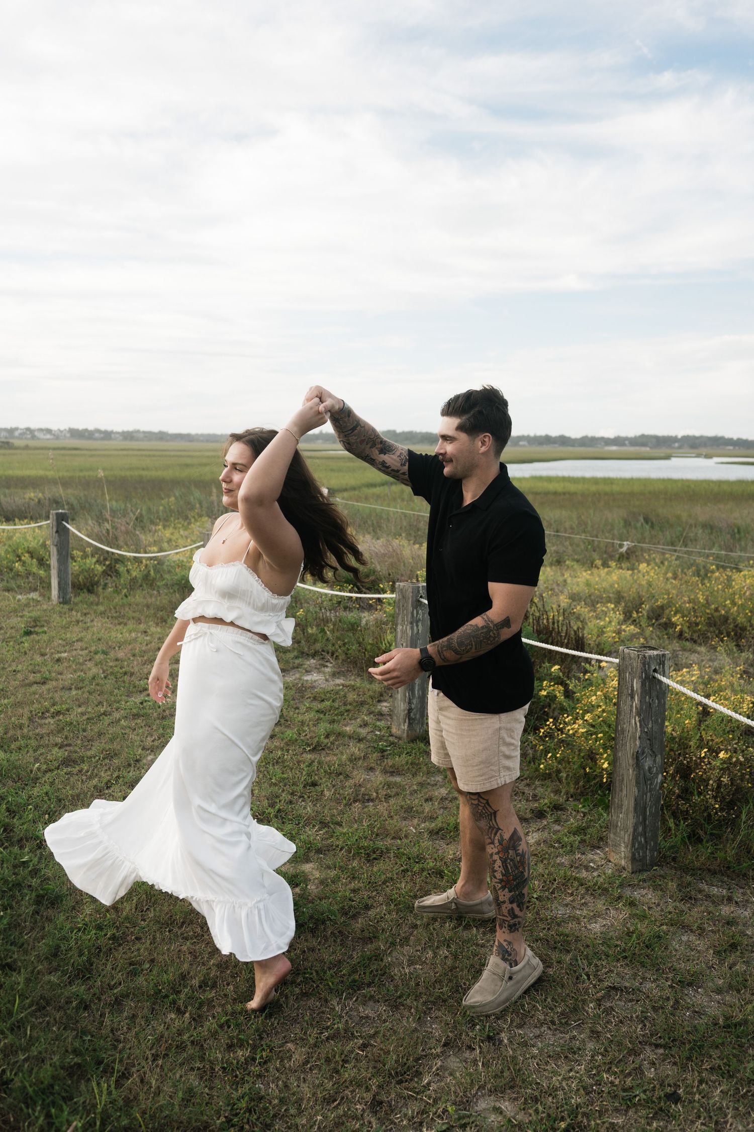 A couple dances together on grassy coastal path with marsh and water views in background.