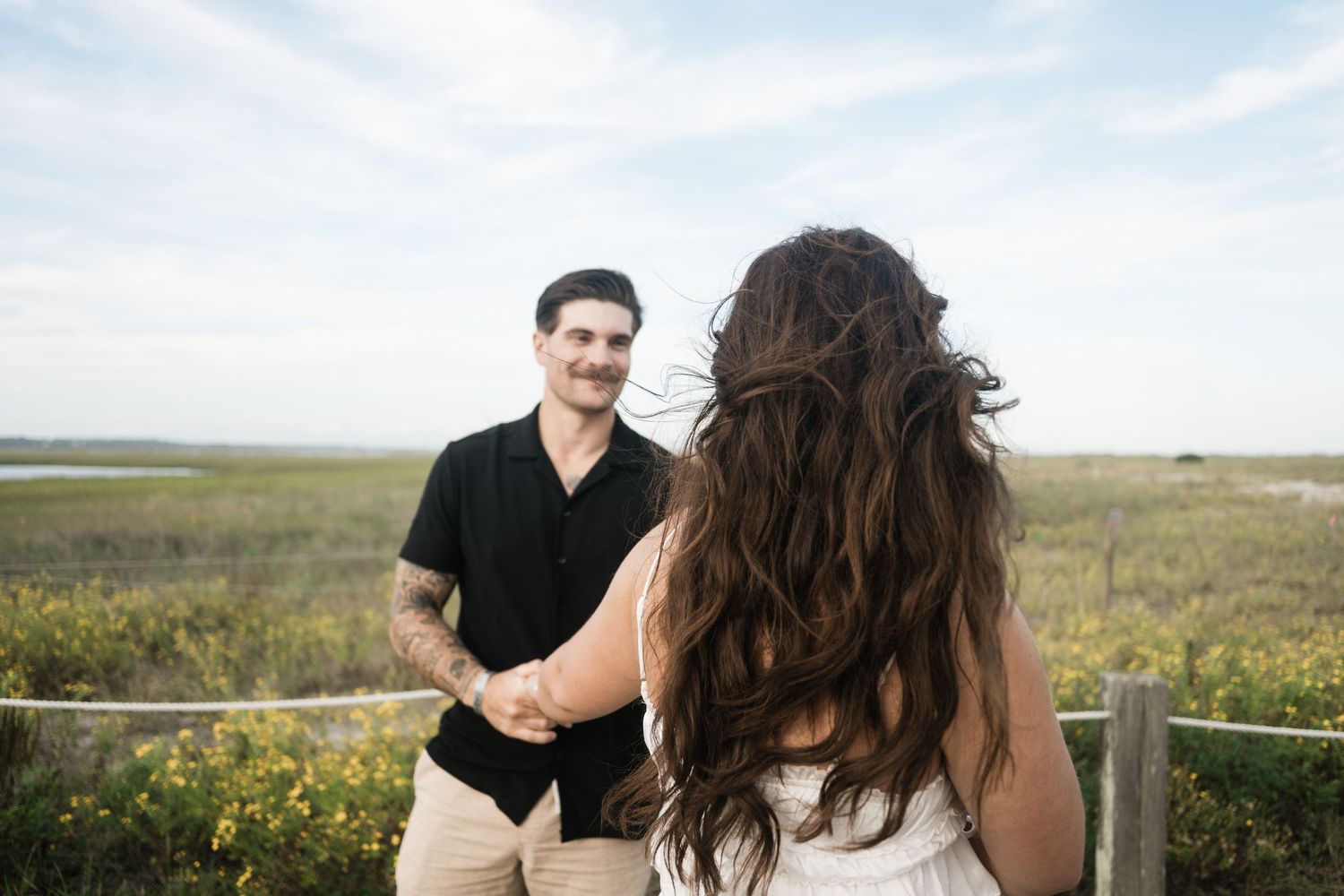 A couple stands together in a scenic field of wildflowers with mountains and blue sky in the background.