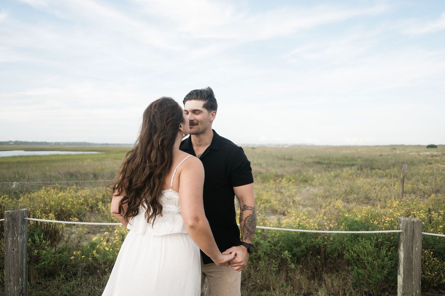 A couple shares an intimate moment together in a scenic coastal field with tall grass and ocean views in the background.