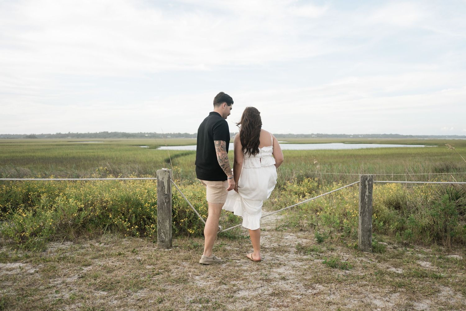 A couple walks hand in hand through scenic coastal grasslands with water and marshland views in the background.