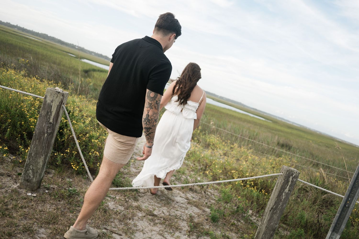 Couple walking along a scenic coastal boardwalk path surrounded by marsh grass and water views.