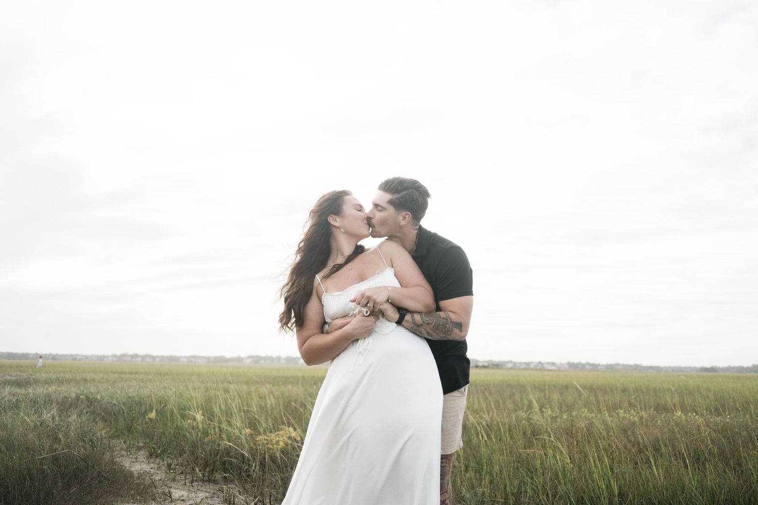 A couple embraces in a romantic moment against a scenic backdrop of tall grass fields and cloudy skies.