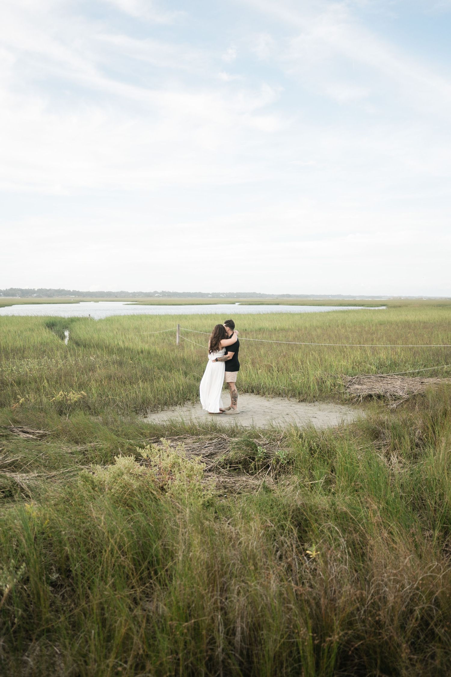 A couple embraces in a scenic coastal marsh landscape with tall grasses and water in the background.