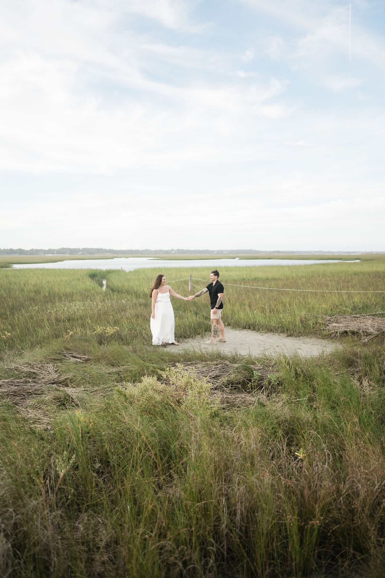 A couple walks along a scenic coastal path with tall grass and water views in the background.