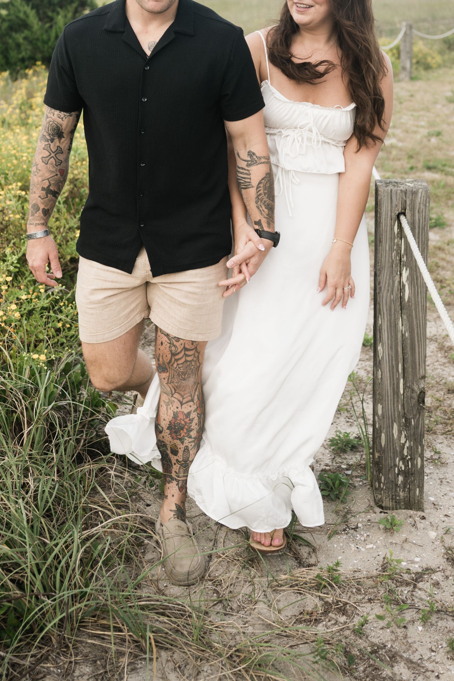 A couple in casual beach attire poses together on sandy ground with rustic wooden fence posts in the background.