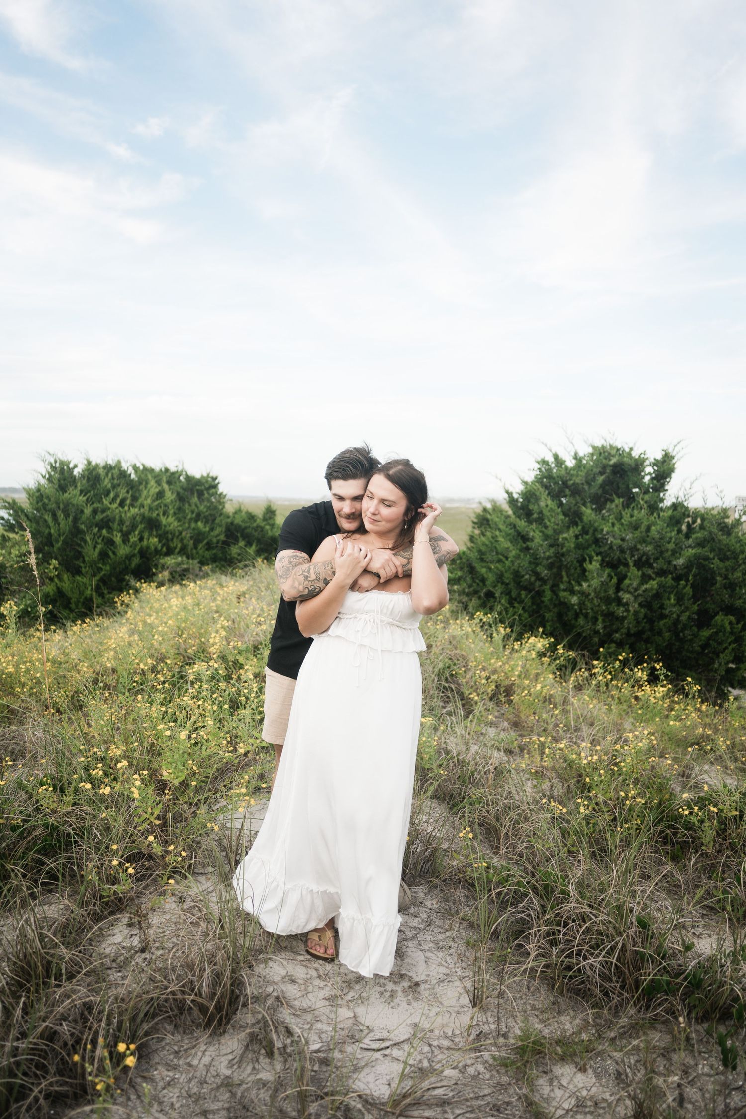A couple in formal attire shares romantic moments on a sandy hill surrounded by yellow wildflowers and greenery.