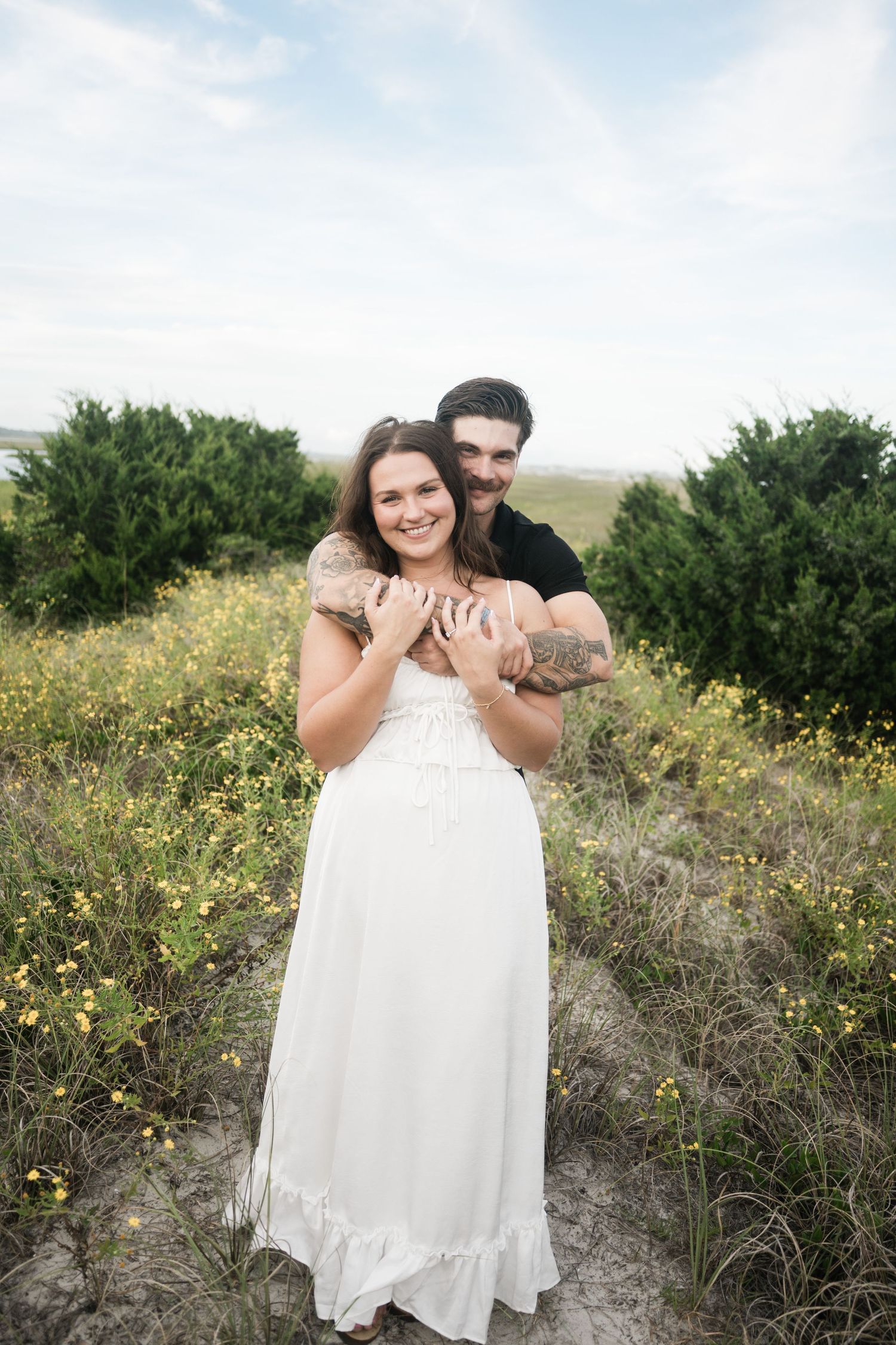 A couple shares a tender moment among yellow wildflowers and coastal vegetation.