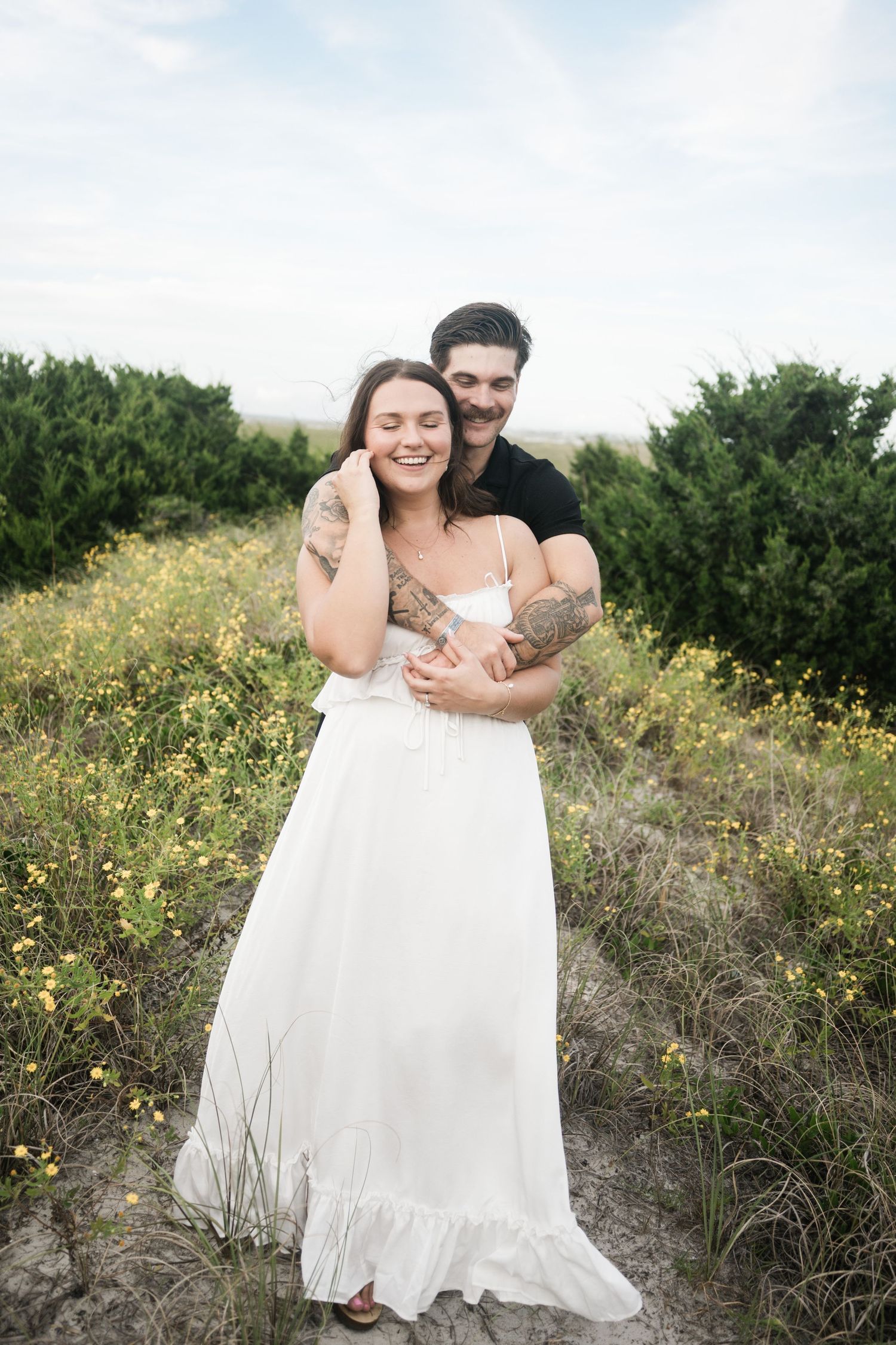 A couple embraces in a field of yellow wildflowers against a backdrop of green bushes during a romantic photoshoot.