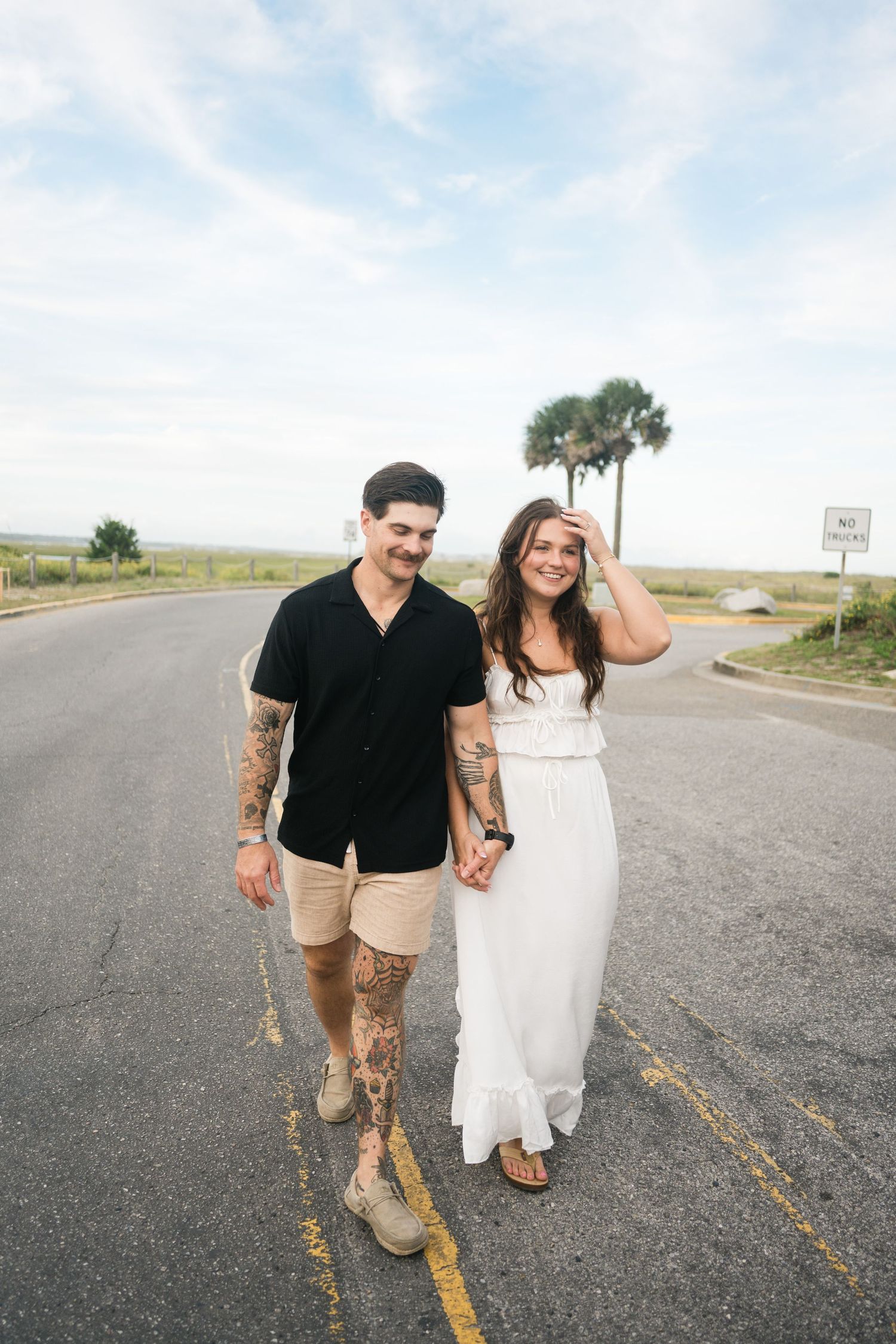 A couple walks hand in hand down a coastal road with palm trees and blue skies in the background.