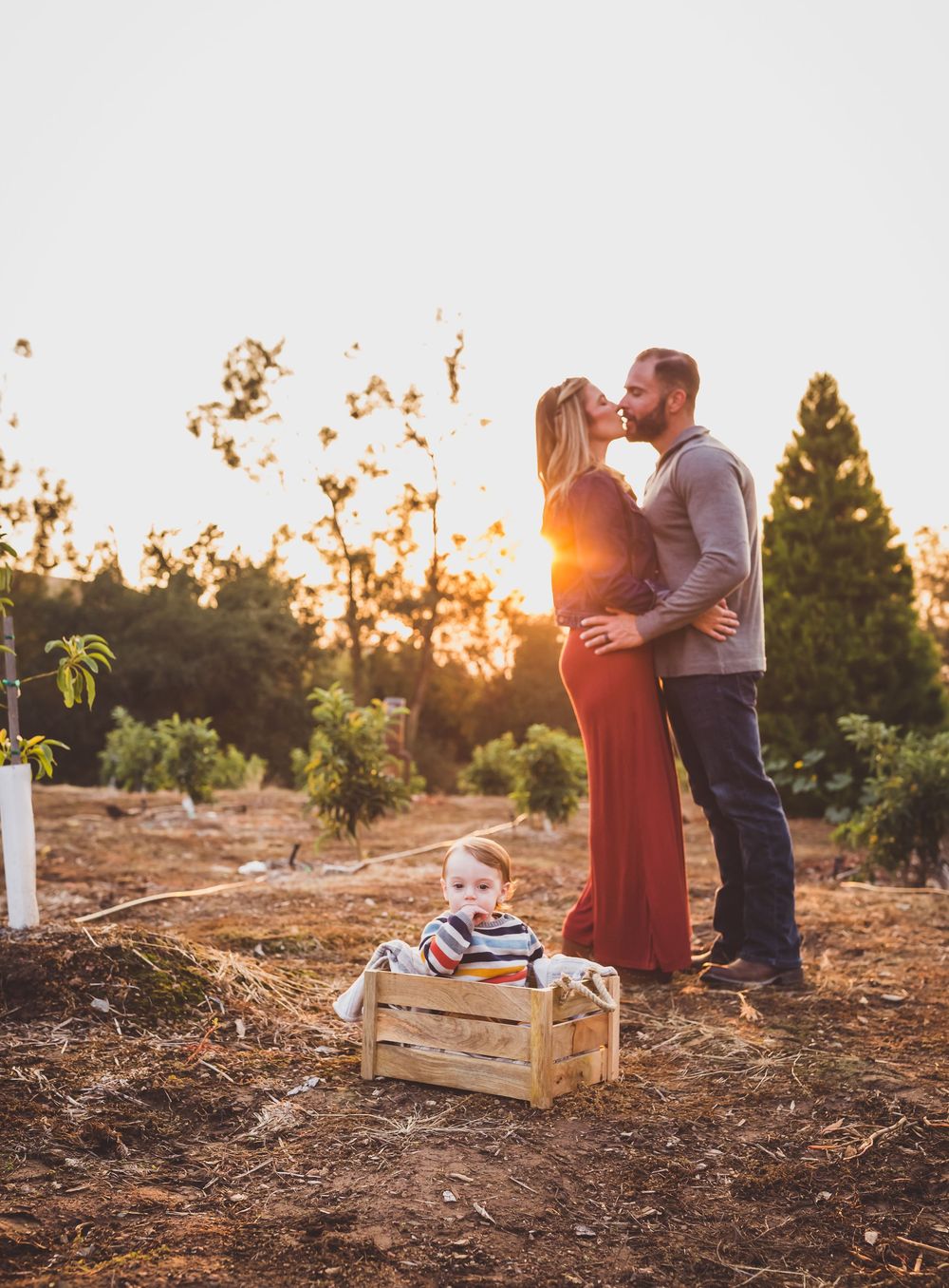 A couple embraces in a rustic outdoor setting at sunset with wooden crates in the background.
