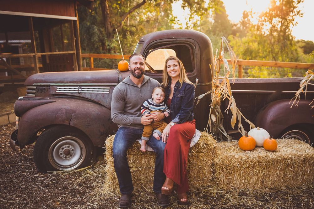 A vintage pickup truck parked in a golden field during sunset creates a warm autumn scene with pumpkins and hay bales.