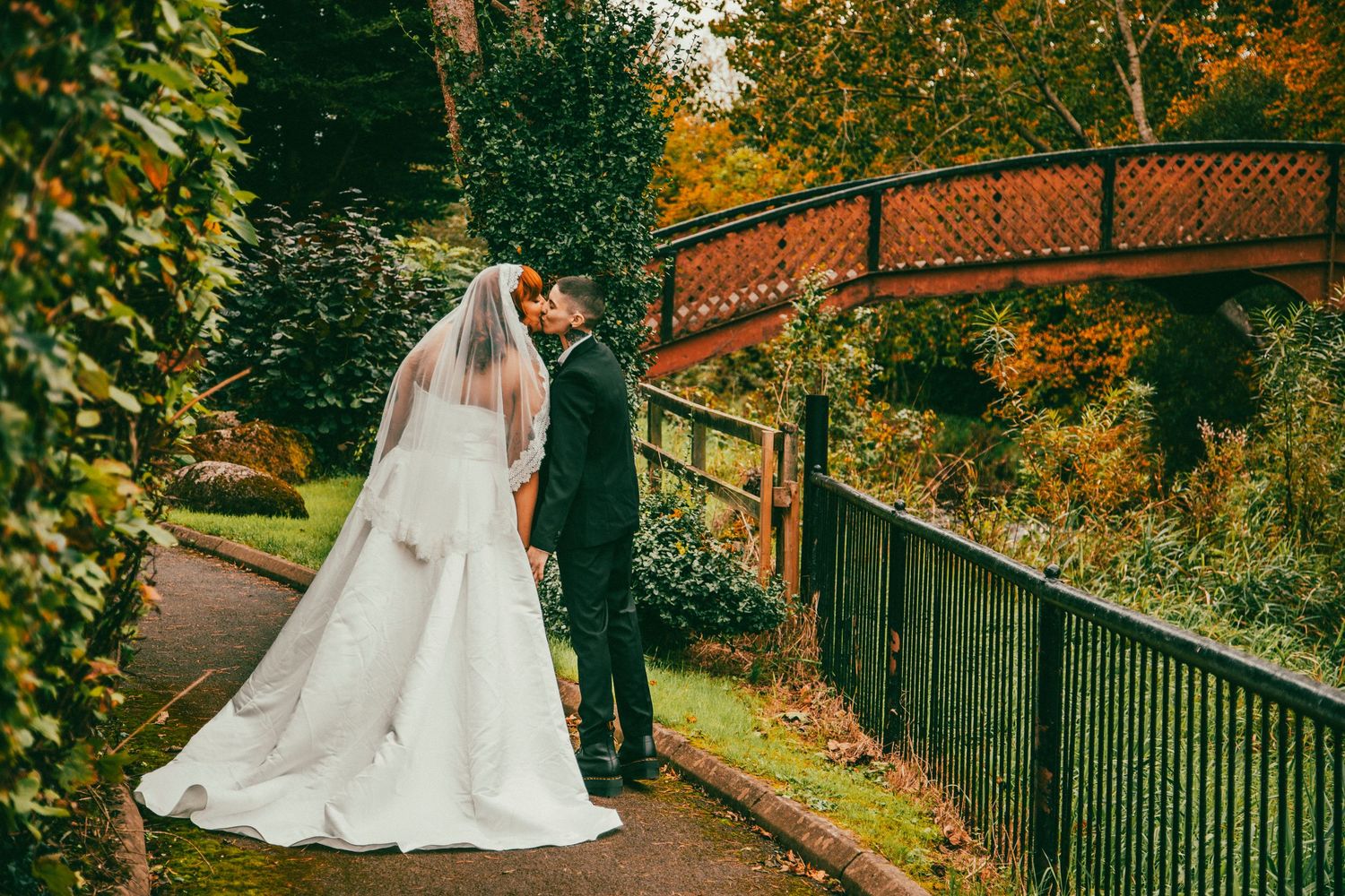 A couple in wedding attire share a romantic moment on a path beneath a rustic bridge surrounded by autumn foliage.