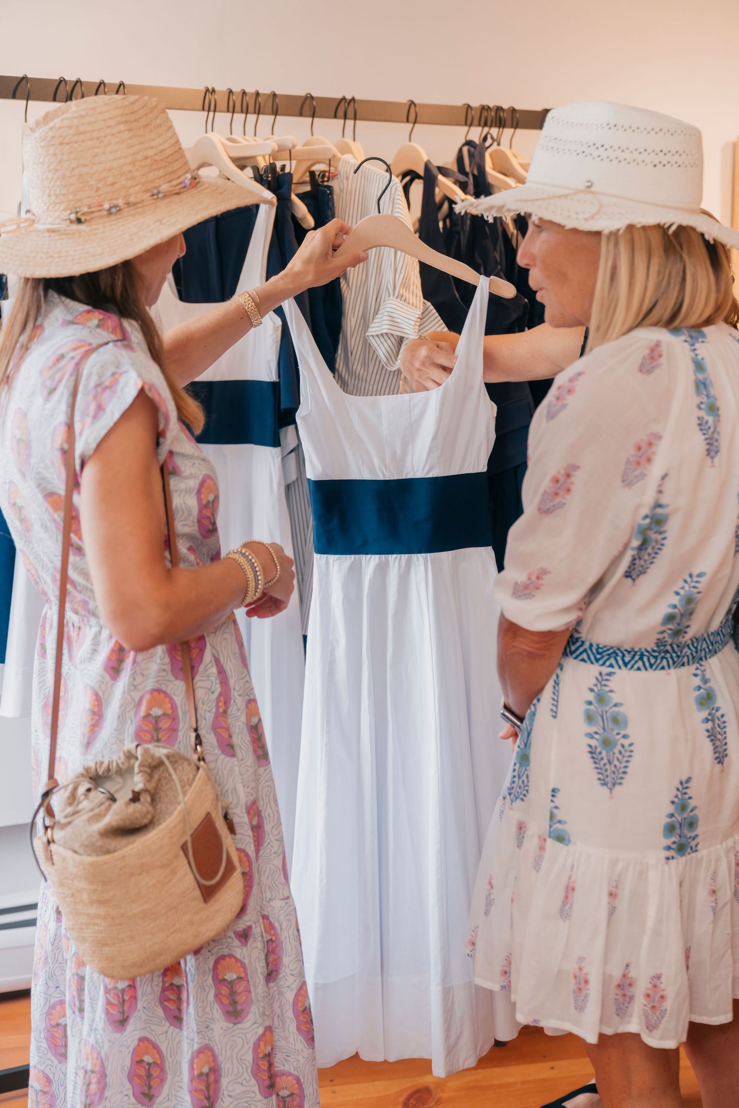 Woman browsing through white and floral dresses on clothing rack while shopping at a boutique store.