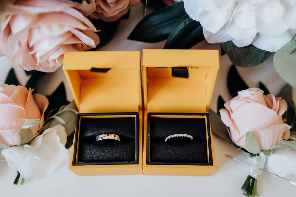 Wedding detail shot of pink roses and engagement rings in golden boxes against white background.