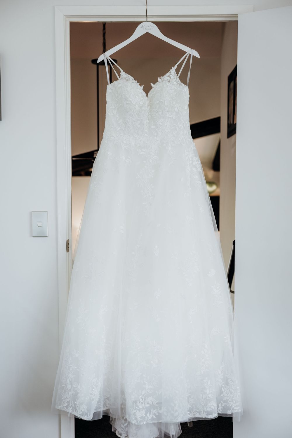 A white lace wedding dress with tulle skirt hangs on a white hanger against a neutral wall.