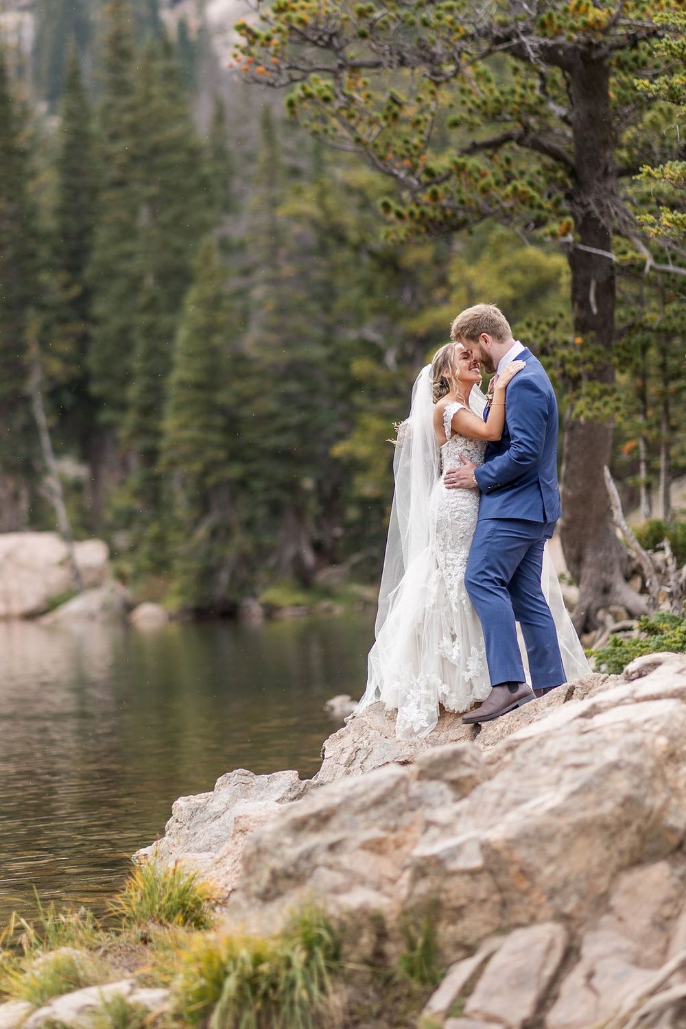 A couple in wedding attire share an intimate moment on rocky terrain by a serene lake surrounded by evergreen trees.