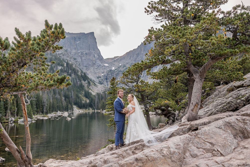 Couple in wedding attire embraces on rocky shore of alpine lake with majestic mountain peaks in background.
