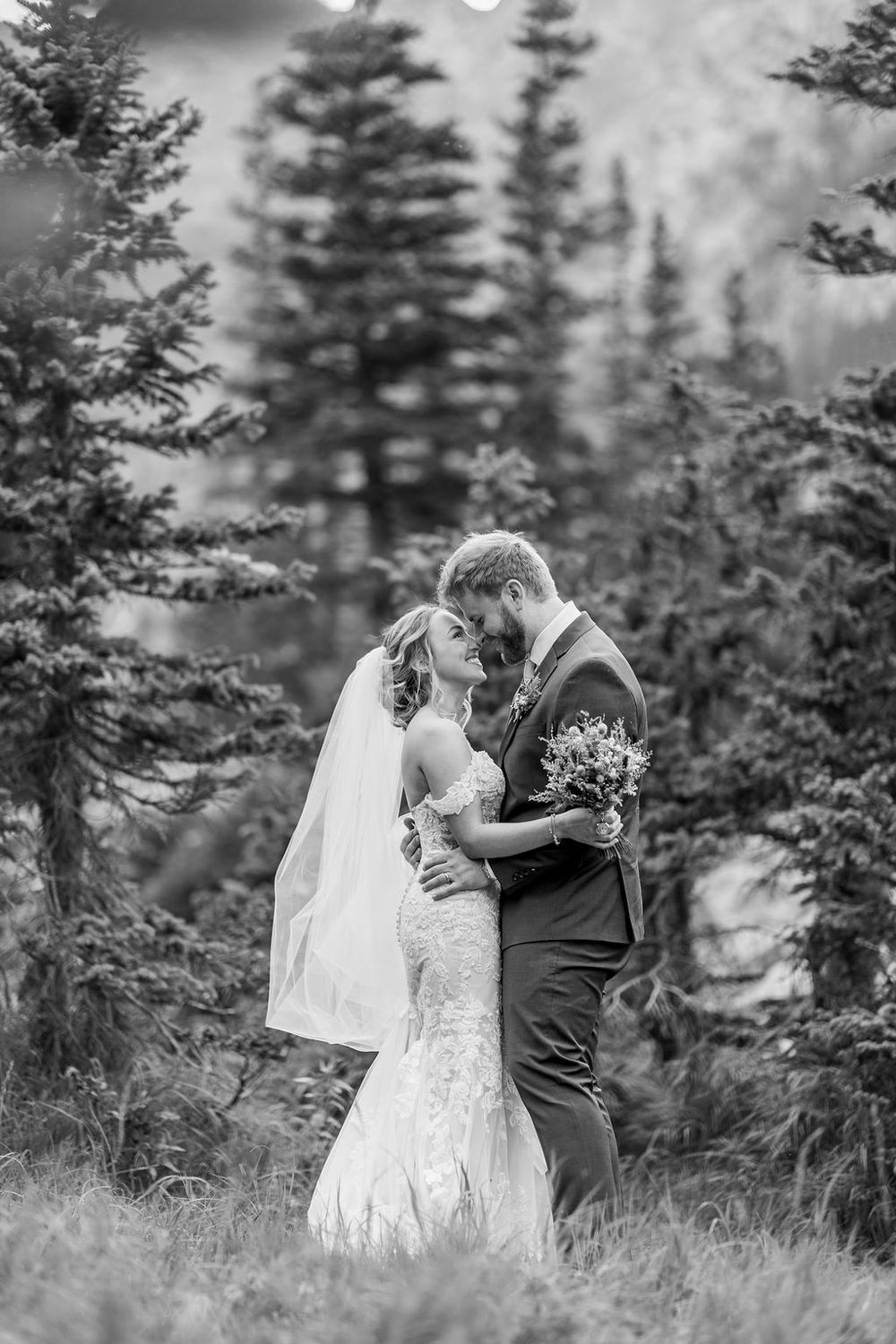 A black and white wedding portrait shows a couple embracing in a mountain setting with evergreen trees in the background.