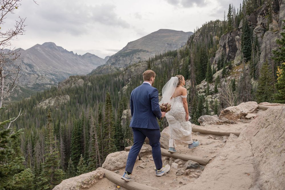 A couple walks along a scenic mountain trail surrounded by evergreen forests and rocky peaks in the distance.