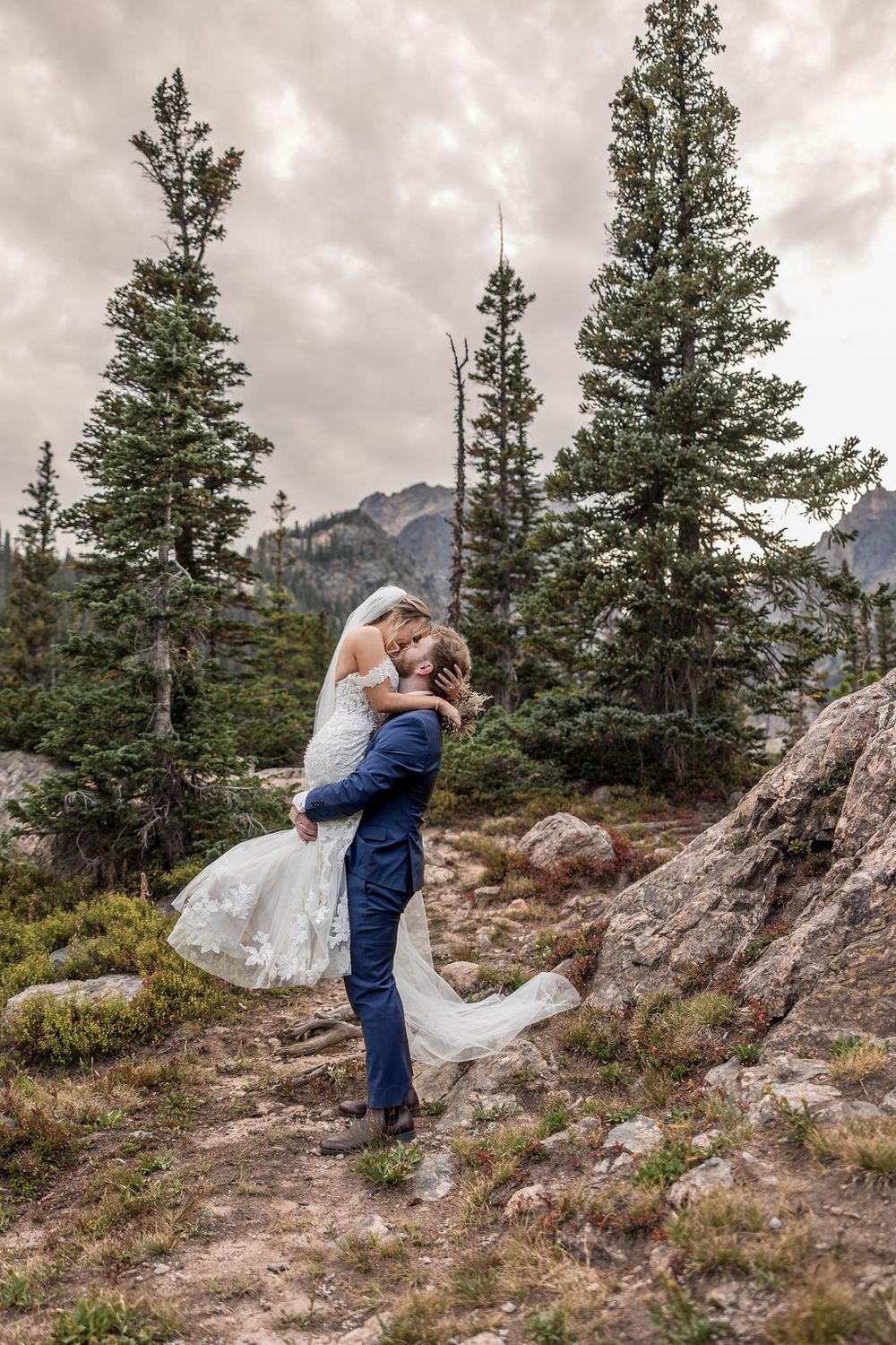 A couple in wedding attire shares romantic moments on a rocky mountain overlook with evergreen trees and cloudy skies.
