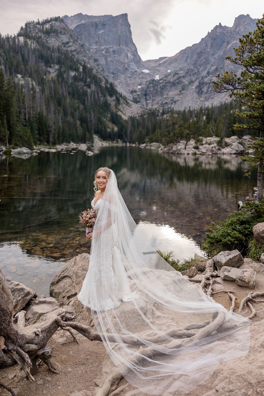 A flowing white wedding dress veil catches the breeze against a stunning mountain lake backdrop in a scenic wilderness setting.