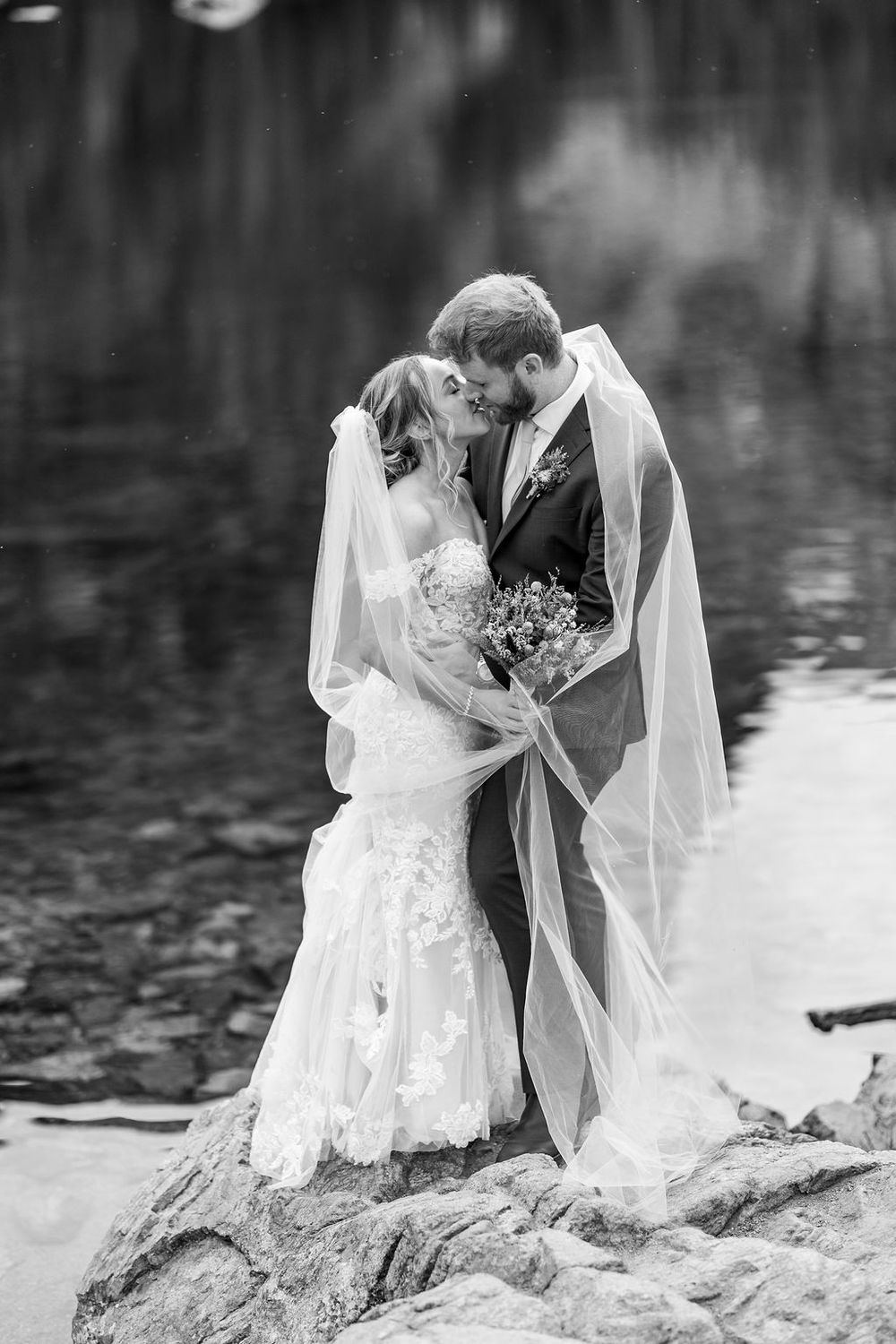 Black and white wedding portraits captured on a rocky lakeside showing intimate moments between newlyweds.