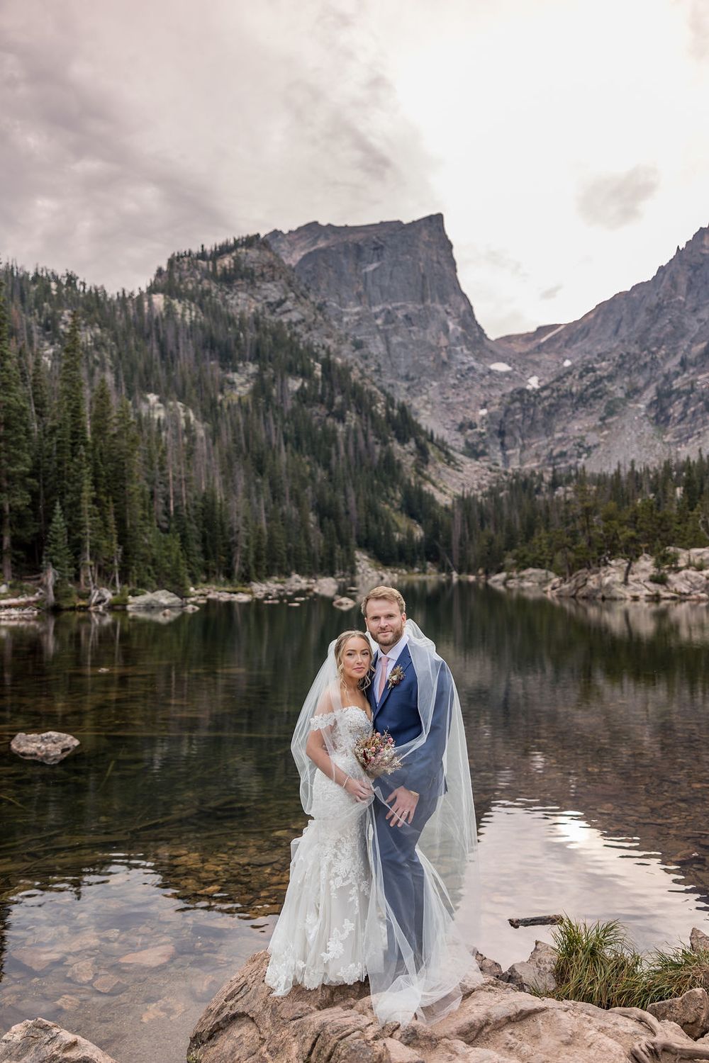 A romantic wedding photo session at a scenic mountain lake with dramatic rocky peaks and evergreen forests in the background.